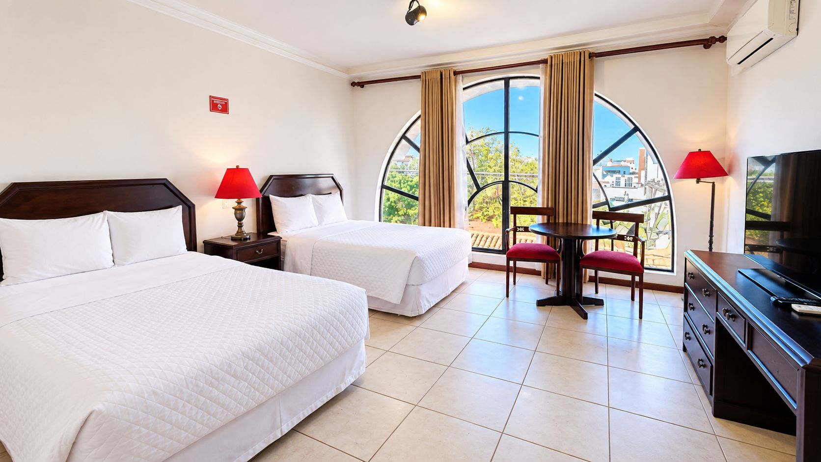 Une chambre d’hôtel avec deux lits, une table à manger avec des chaises, un téléviseur et une grande fenêtre avec des rideaux.