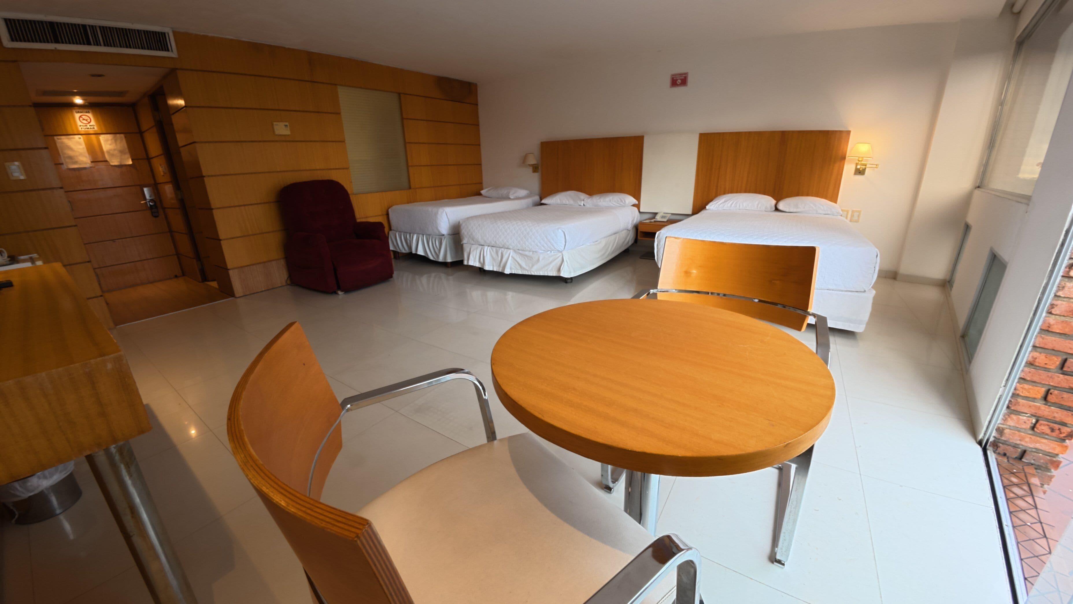A hotel room with a round wooden table, two single beds, a red chair, and a wooden wall.