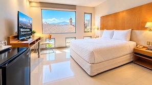 Habitació d'hotel amb un llit, una taula de treball, un televisor i un gran finestral amb vista a la ciutat.