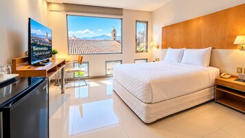 Habitación de hotel con cama, escritorio, televisor y una ventana grande con vistas a la ciudad.
