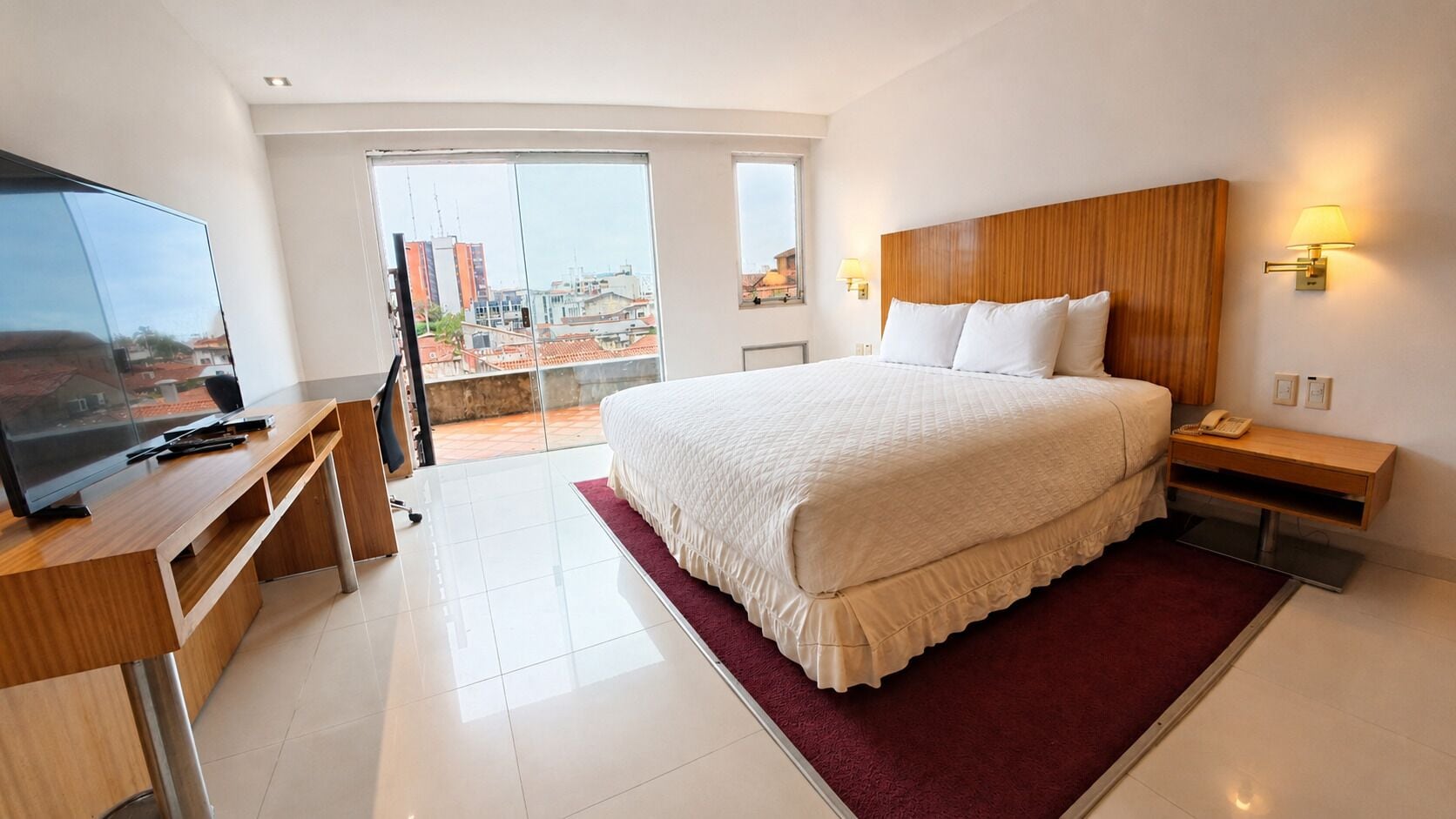 A hotel room with a large bed, a desk with a TV, and a view of the city through the window.