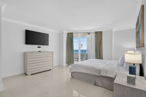 Una habitación de hotel con una cama, un televisor montado en la pared, una mesita de noche con lámpara y vistas al mar a través de las puertas del balcón.