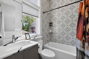 Un baño moderno con un lavamanos blanco, un espejo y una bañera. Hay artículos de aseo en un estante y una fotografía enmarcada sobre el mostrador.