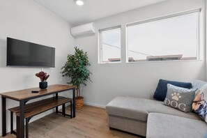 A modern living room with a flat-screen TV, a wooden table with a bench, a potted plant, a grey sofa with a dog-themed pillow, and a large window with a view of a building. - Stunning water view, pet friendly, luxury 3-bedroom townhome with AC in Seattle (Seattle)