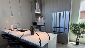 Moderni keittiö, jossa on keskeinen saareke, baarituolit, ruostumattomasta teräksestä valmistetut kodinkoneet, riippuvalaisimet ja ruukussa oleva kasvi.