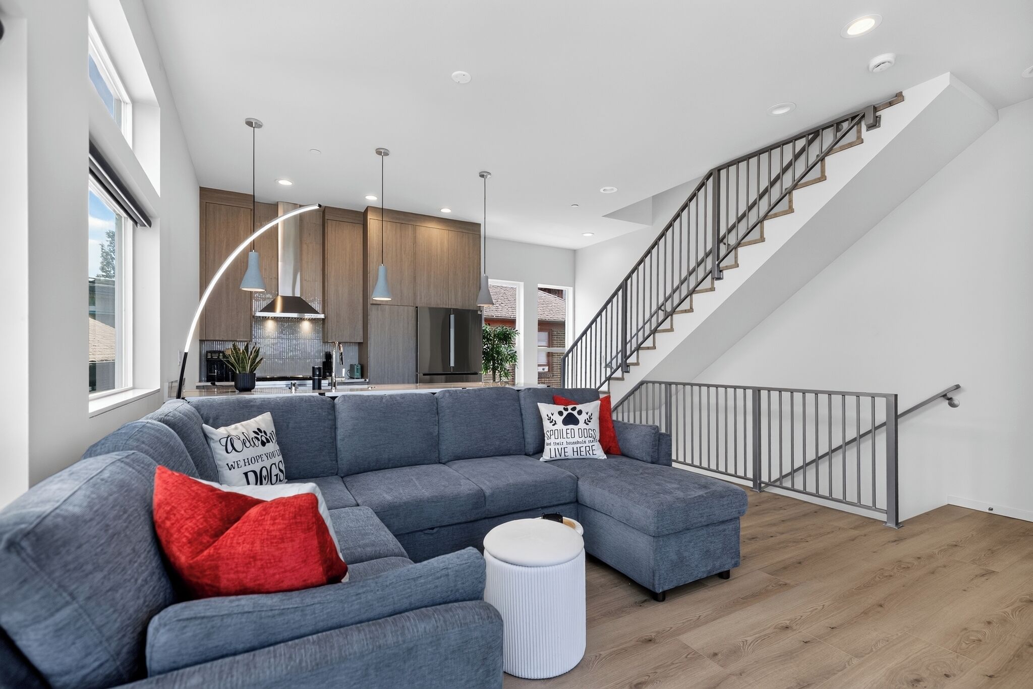 Ein modernes Wohnzimmer mit einem grauen Ecksofa, einem runden Hocker und einer Treppe mit Metallgeländer.