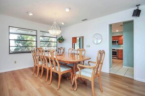 A dining area with a round wooden table and chairs, a chandelier, and a view of the kitchen through an open doorway. - Waterside Retreat with Pool, Steps to Lighthouse (Sanibel)