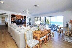 A spacious living room with a large sofa, a wooden dining table, and a view of the ocean through large windows. - Waterside Retreat with Pool, Steps to Lighthouse (Sanibel)