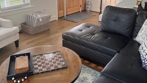 A living room with a black leather sofa, a wooden coffee table with chess pieces, a white armchair, and a large window with blinds.
