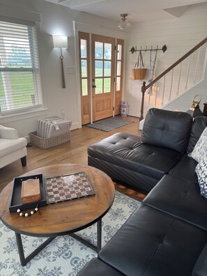 A living room with a black leather sofa, a wooden coffee table with chess pieces, a white armchair, and a large window with blinds.
