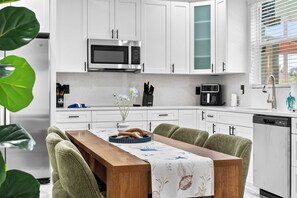 Una cocina moderna con armarios blancos, un microondas de acero inoxidable, una cafetera y un comedor con mesa de madera y sillas tapizadas de color verde.
