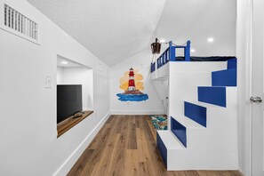 Una habitación con suelo de madera, un televisor sobre un mueble, una escalera con peldaños azules y un mural en la pared con un faro.