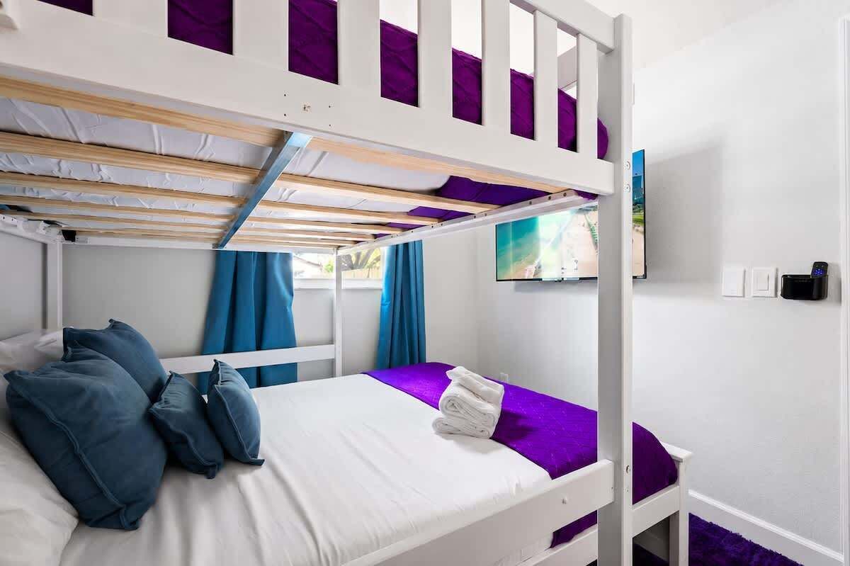 Une chambre avec un lit superposé, un cadre blanc, une literie violette et un téléviseur fixé au mur.