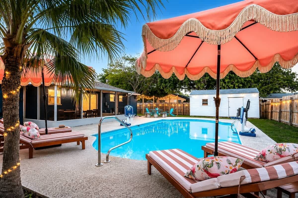 Ein Swimmingpool mit Liegestühlen und einem Sonnenschirm, umgeben von einem Haus und Bäumen.