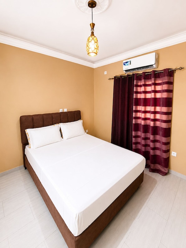 A bedroom with a bed, a ceiling light, an air conditioning unit, and curtains.