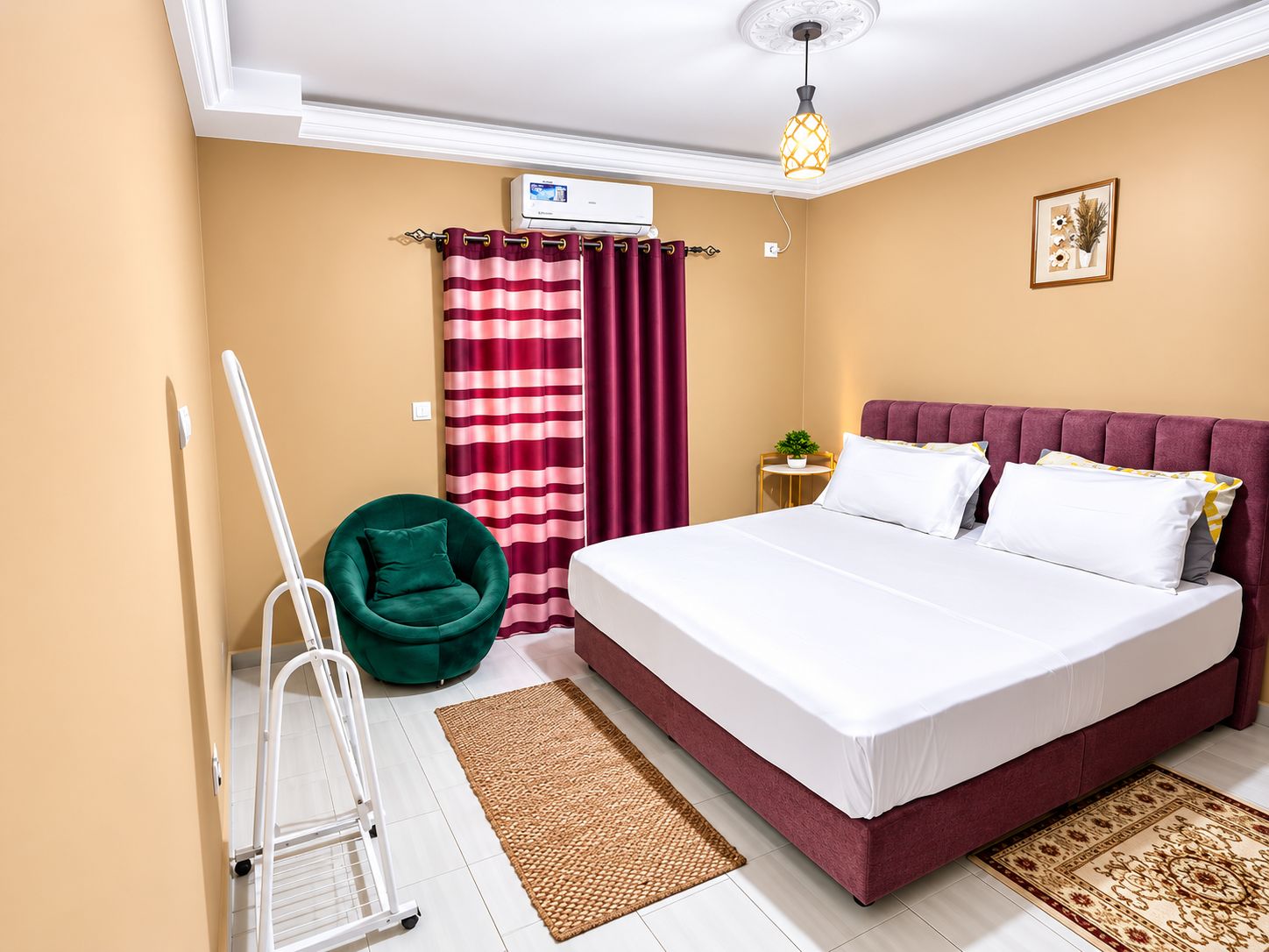 Une chambre d’hôtel avec un lit, un fauteuil vert, une petite table avec une plante, un rideau à rayures rouges et blanches, et un luminaire au plafond.