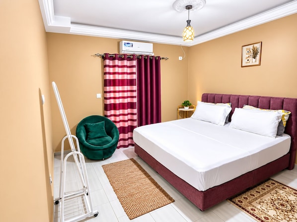 A hotel room with a bed, a green chair, a small table with a plant, a red and white striped curtain, and a ceiling light fixture.