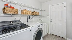 A laundry area with a washing machine, dryer, and shelves with baskets and cleaning supplies. - Spacious 4-bedroom house in serene Pensacola with private pool (Pensacola)
