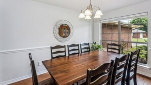 A dining room with a wooden table and chairs, a chandelier, a mirror, and a large window with a view of a brick building and greenery. - Spacious 4-bedroom house in serene Pensacola with private pool (Pensacola)
