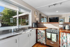A kitchen with a stainless steel sink, a stove, and a window with a view of trees. - Coastal Hideaway - Waipu Cove Holiday Home (Waipu)