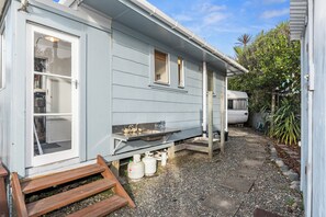 A small, single-story house with a front porch, a small table, and a window. - Coastal Hideaway - Waipu Cove Holiday Home (Waipu)