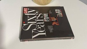 Un libro titulado 'LIFE SIXTY Years' sobre una superficie blanca, con una planta en maceta al fondo.