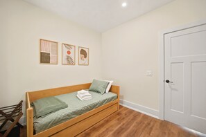 Un lit simple avec une couverture et des oreillers verts, une tête de lit en bois, une petite chaise en bois et une porte sur la droite.