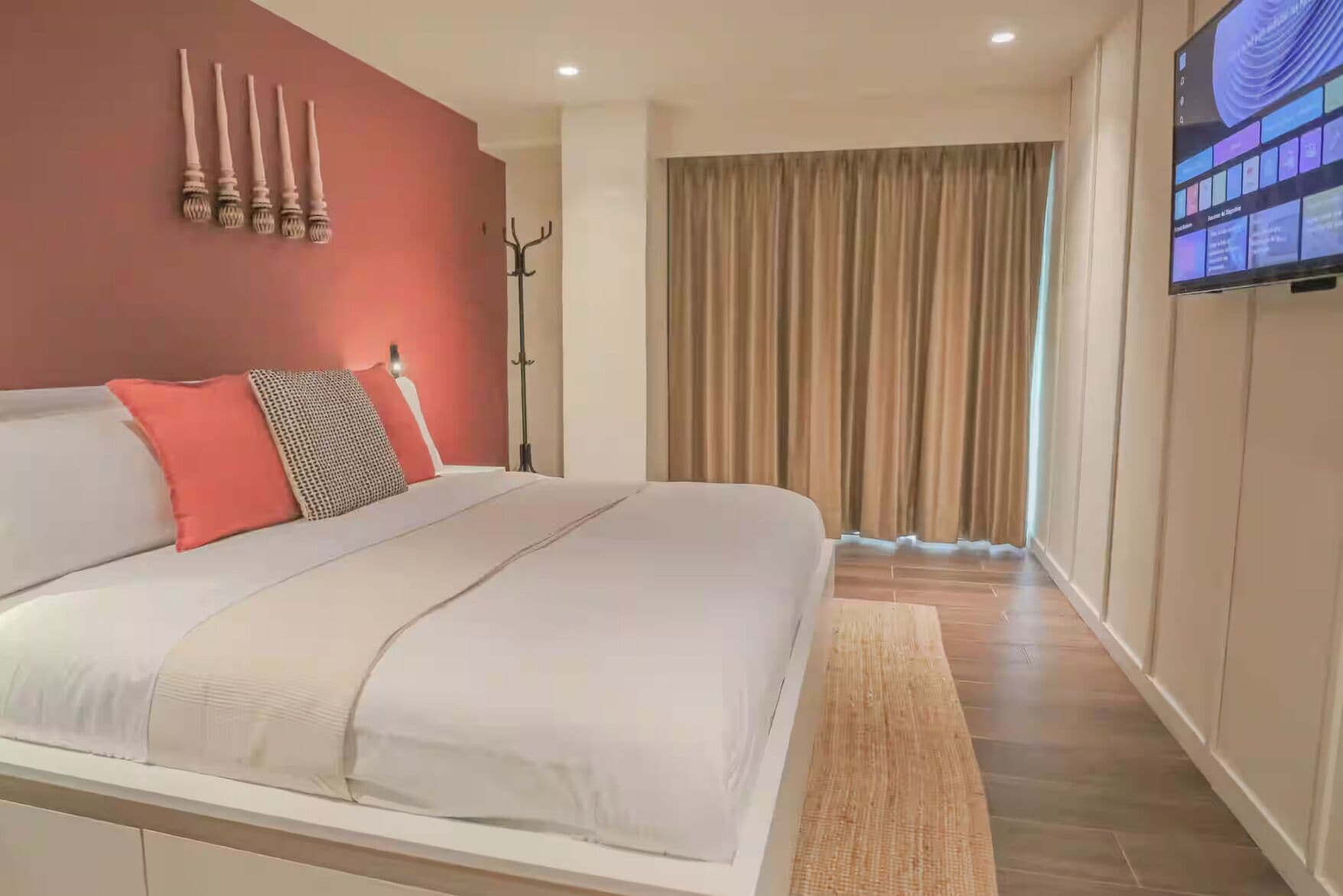 A modern hotel room with a large bed, a flat-screen TV, a coat rack, and a view of the cityscape through the window.