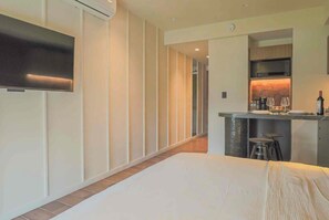 Habitación de hotel con cama, televisor montado en la pared, kitchenette con microondas y copas de vino, y una pequeña zona de bar.