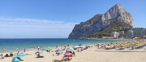 A beach with umbrellas, sun loungers, and a rocky cliff in the background.