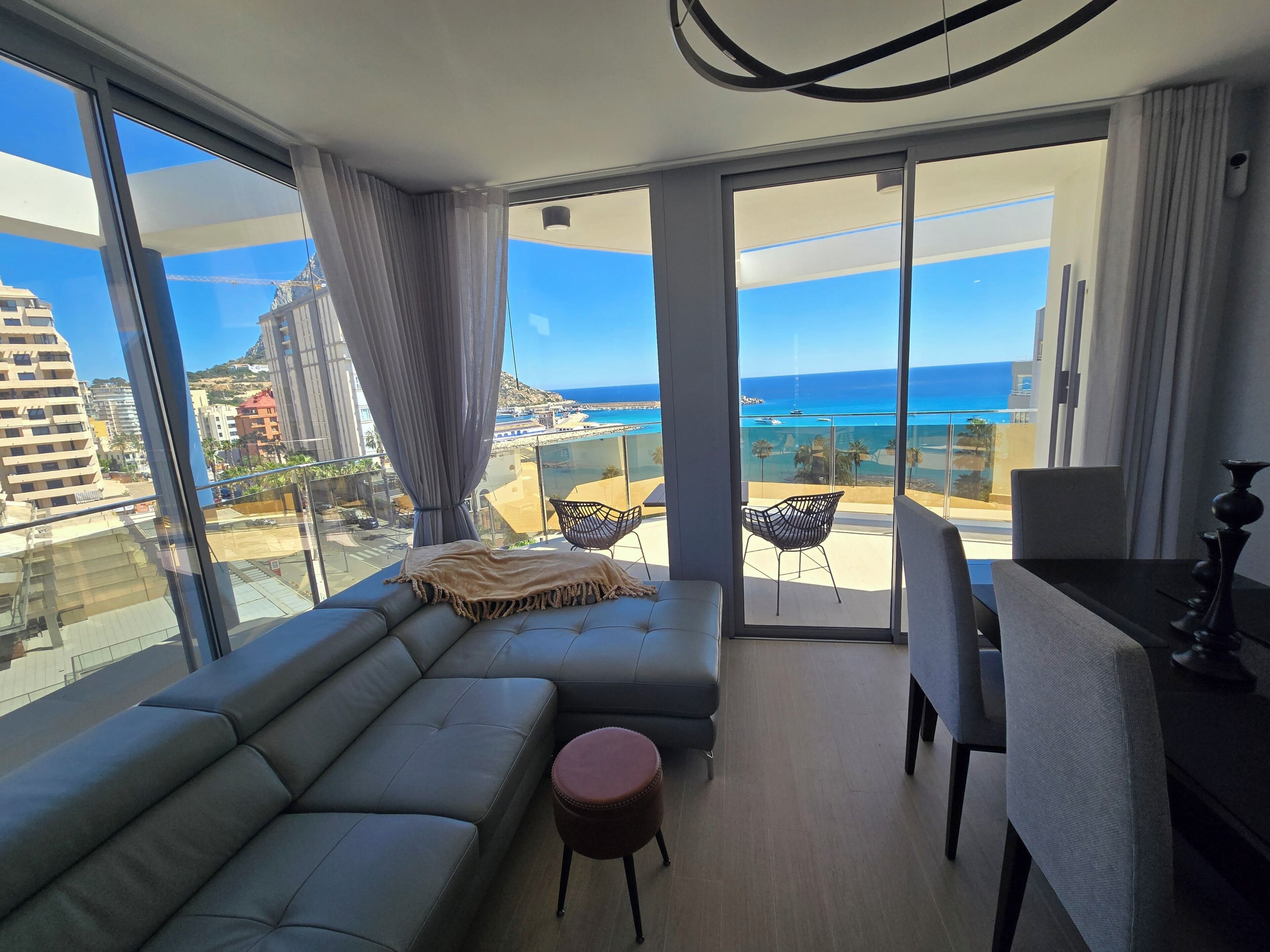 A modern living room with a large sofa, a small round ottoman, a dining table with chairs, and a view of the ocean and buildings.