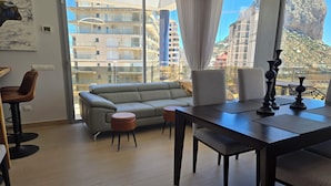 Een moderne woonkamer met een bank, een eettafel en uitzicht op een stadsgezicht door grote ramen.