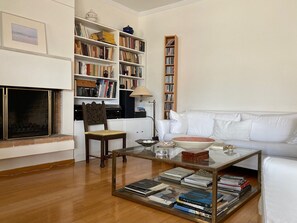Ein Wohnzimmer mit Kamin, einem weißen Sofa, einem Holzregal und einem Glastisch mit Büchern.