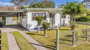 A single-story building with a covered outdoor area, a paved driveway, and a fenced yard with small trees. - Camel Rock Cottage - Beauty Point 33 (Wallaga Lake)
