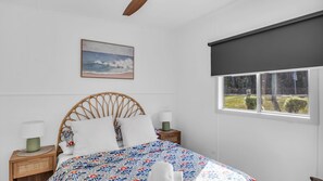 A bedroom with a bed, a nightstand, a lamp, a window with a view of the outdoors, and a painting on the wall. - Camel Rock Cottage - Beauty Point 33 (Wallaga Lake)