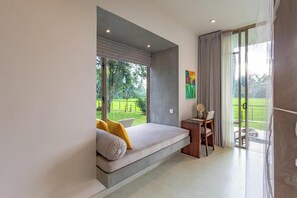 A modern bedroom with a built-in sofa bed, a small table with chairs, and a view of a green garden through large windows. - ONA Garden Villa: Double Room with Terrace (Wanchawala)