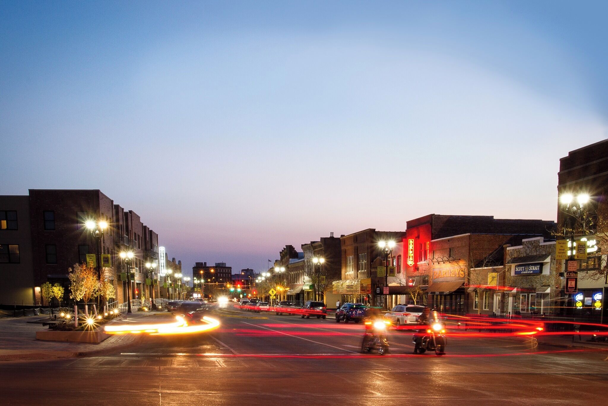 A city street at dusk with illuminated buildings and moving traffic.
