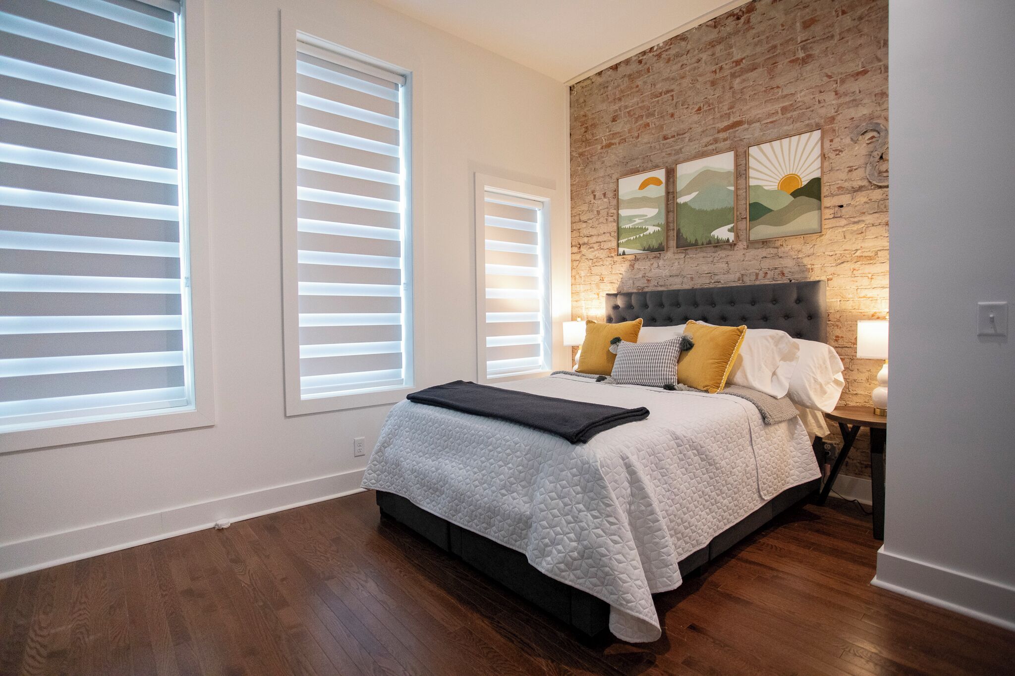 A bedroom with a bed, two windows with blinds, a brick wall, and a nightstand with a lamp.