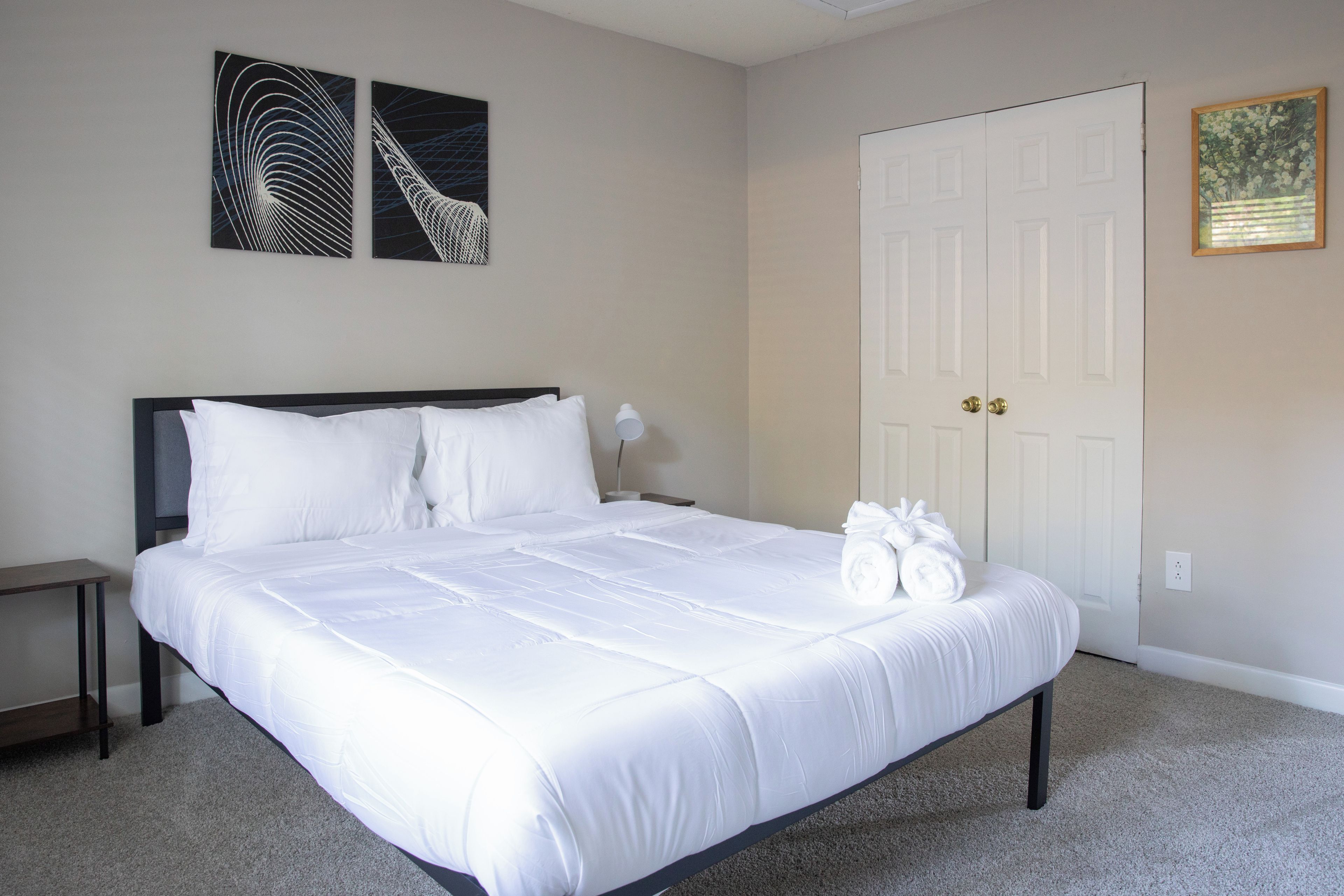 A neatly made bed with white linens, a nightstand with a lamp, and two wall art pieces.