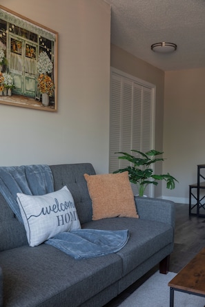 Una sala de estar con un sofá gris, un cojín con la frase 'bienvenido a casa', una planta en maceta y un cuadro en la pared.