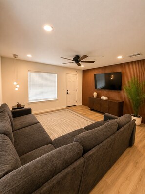 A modern living room with a sectional sofa, a flat-screen TV mounted on a wooden wall, a ceiling fan, and recessed lighting.