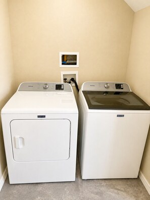 Two Maytag front-loading washing machines in a laundry room with beige walls and a small framed picture on the wall.