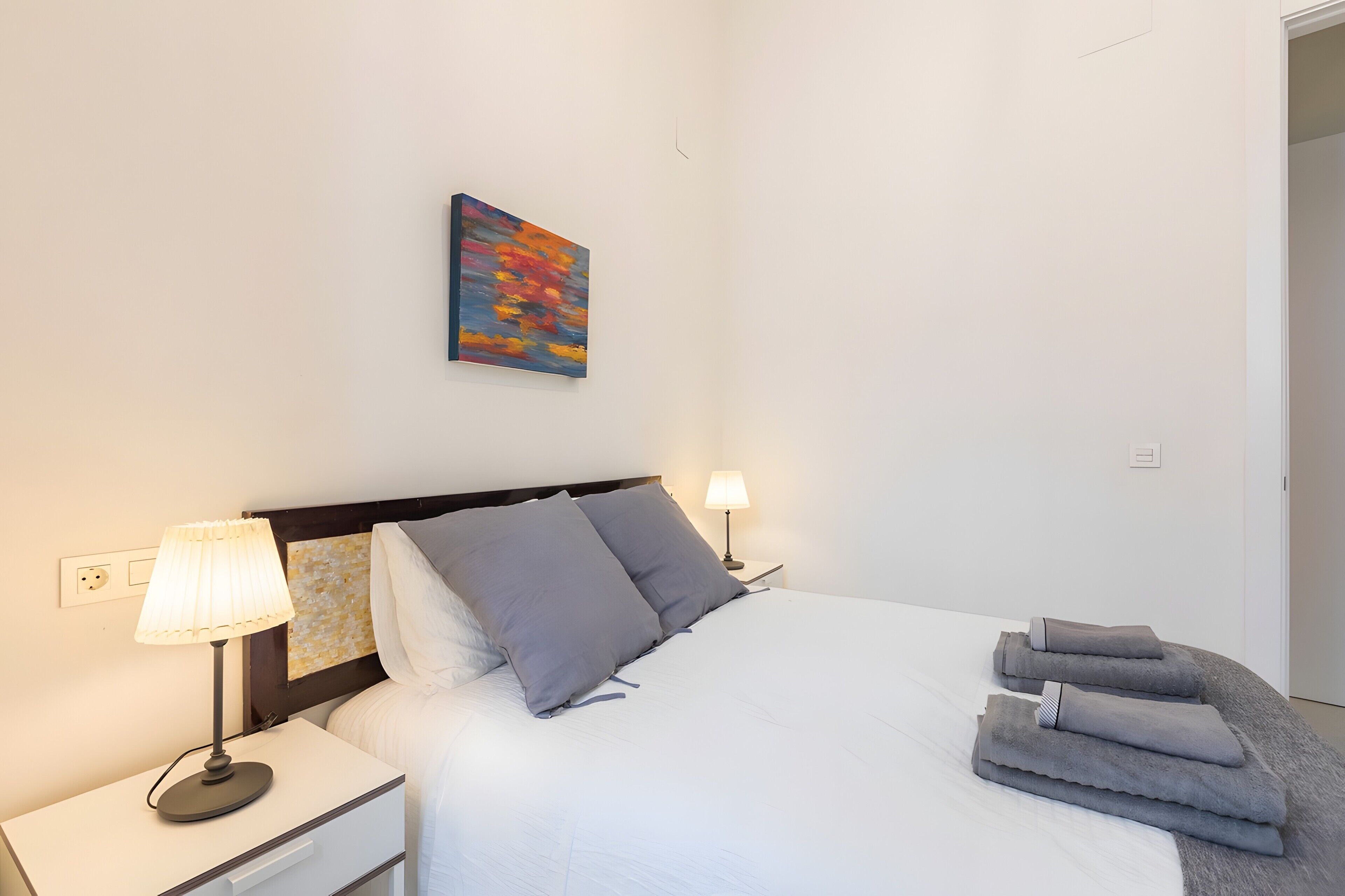 Uma cama bem arrumada com almofadas e um cobertor, uma luminária de cabeceira, um quadro na parede e uma pilha de toalhas no chão.