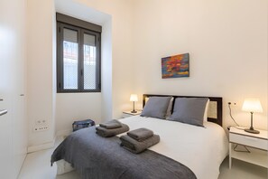A hotel room with a bed, two bedside tables with lamps, a painting on the wall, and a window with a view. - Cozybeautiful Apartment with Wi-Fi and Air Conditioning (Seville)