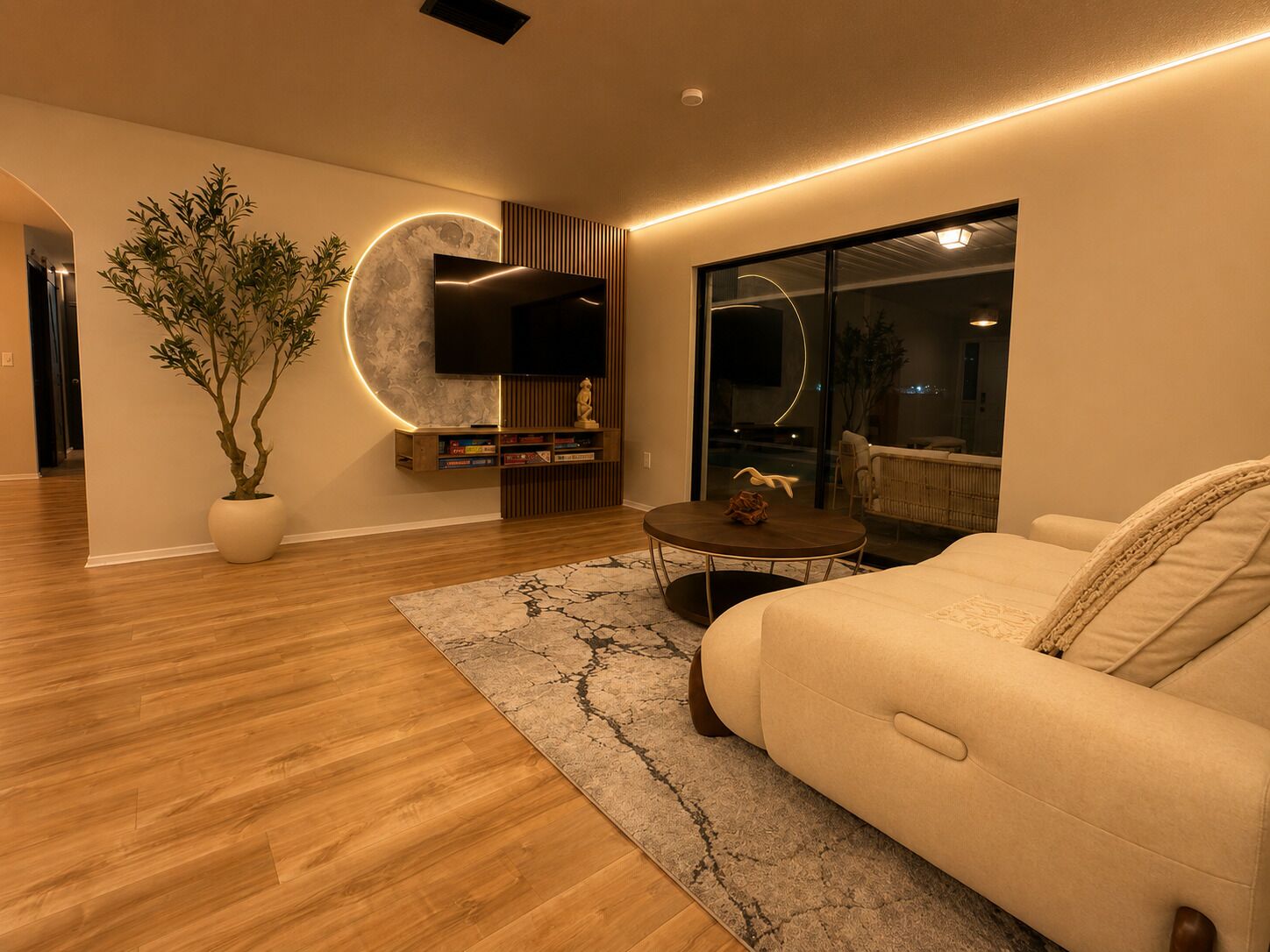A modern living room with a sofa, a coffee table, a TV mounted on a wall, a potted plant, and a view through a glass door.