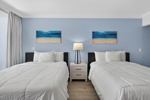 A hotel room with two beds, a nightstand with a lamp, and two wall-mounted paintings of a beach scene. - Blue Horizon (Fort Lauderdale)