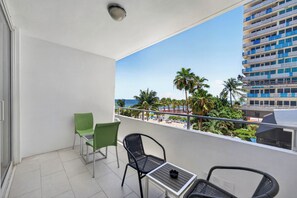 A balcony with a view of the beach and palm trees, a table, two chairs, and a ceiling light. - Blue Horizon (Fort Lauderdale)