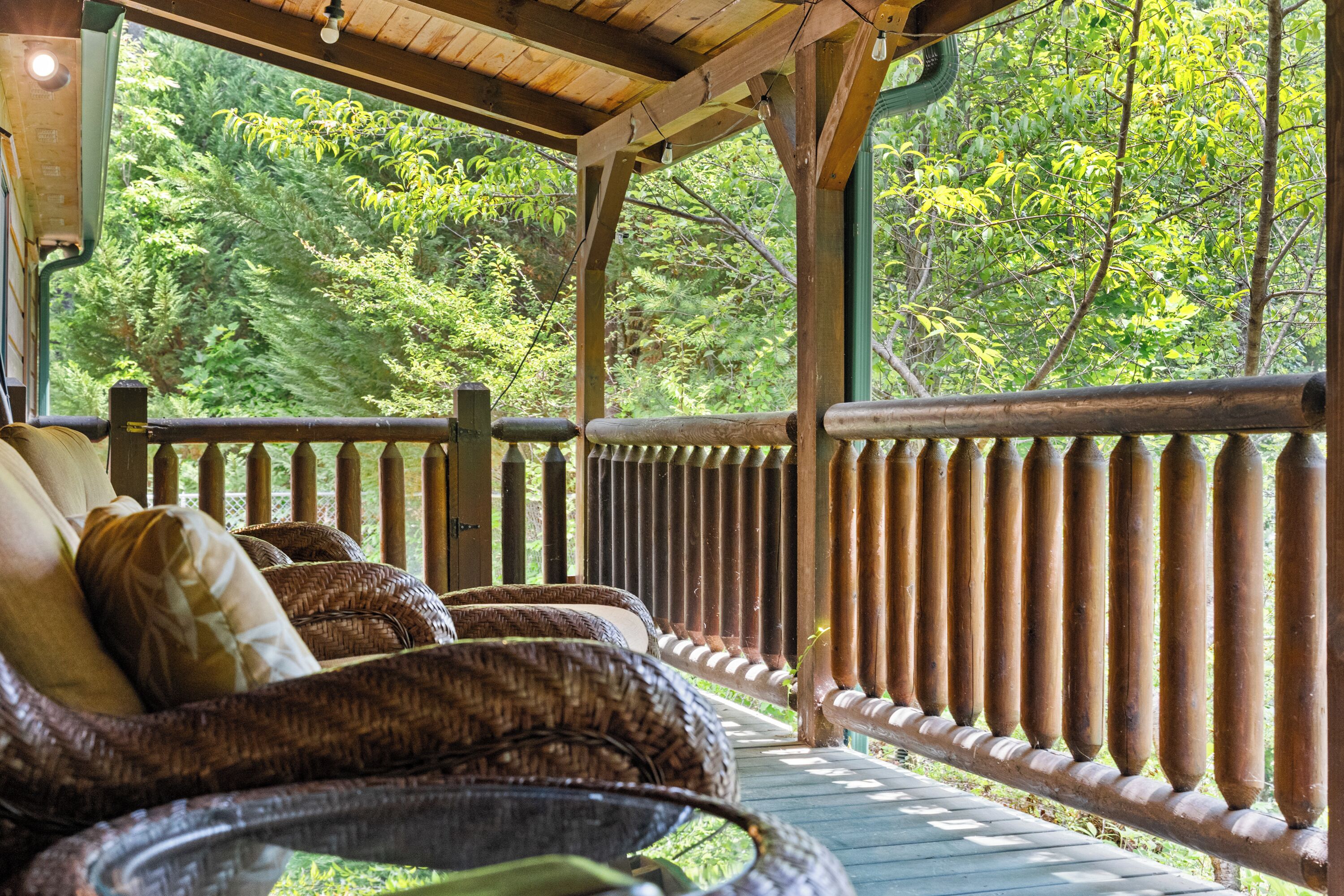 A covered outdoor seating area with wicker furniture and a wooden railing overlooking a forested area.