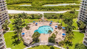 A hotel with a central swimming pool surrounded by lounge areas, palm trees, and a view of the ocean. - Beachfront Paradise - Stunning Gulf Views - Luxury 17th Floor Condo (Marco Island)
