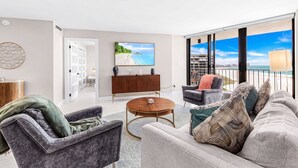 A modern living room with a sofa, armchairs, a coffee table, and a TV. There is a balcony with a view of the beach and buildings. - Beachfront Paradise - Stunning Gulf Views - Luxury 17th Floor Condo (Marco Island)
